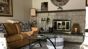 Un salón acogedor con un sofá de cuero marrón, una mesa de centro de vidrio y cojines estampados. Un espejo decorativo grande cuelga de la pared, y se aprecia una chimenea con un revestimiento de piedra.