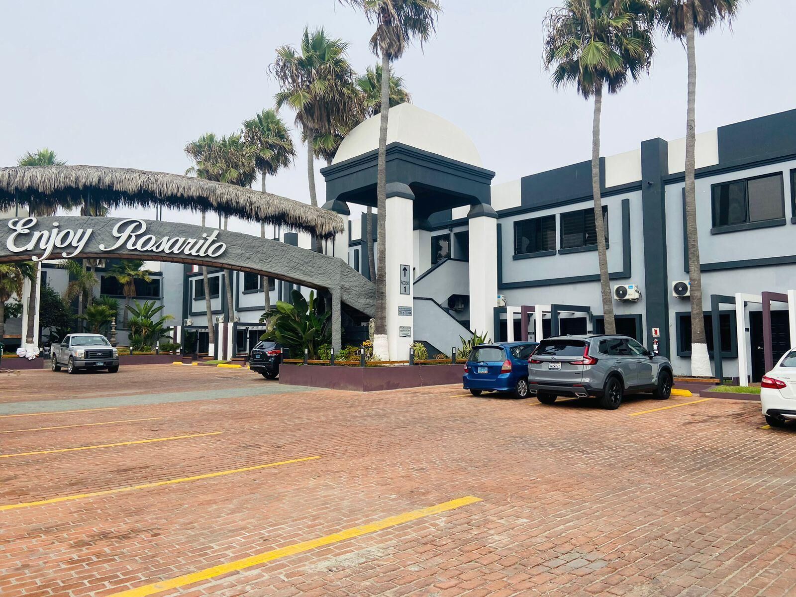 A hotel entrance with a sign reading "Enjoy Rosarito," featuring a modern building with a thatched roof entrance, palm trees, and parked cars.