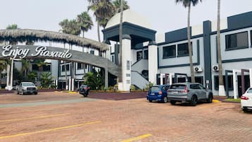 Entrada de hotel con un cartel que dice "Enjoy Rosarito", que presenta un edificio moderno con techo de paja, palmeras y coches estacionados.