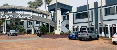 A hotel entrance with a sign reading "Enjoy Rosarito," featuring a modern building with a thatched roof entrance, palm trees, and parked cars.