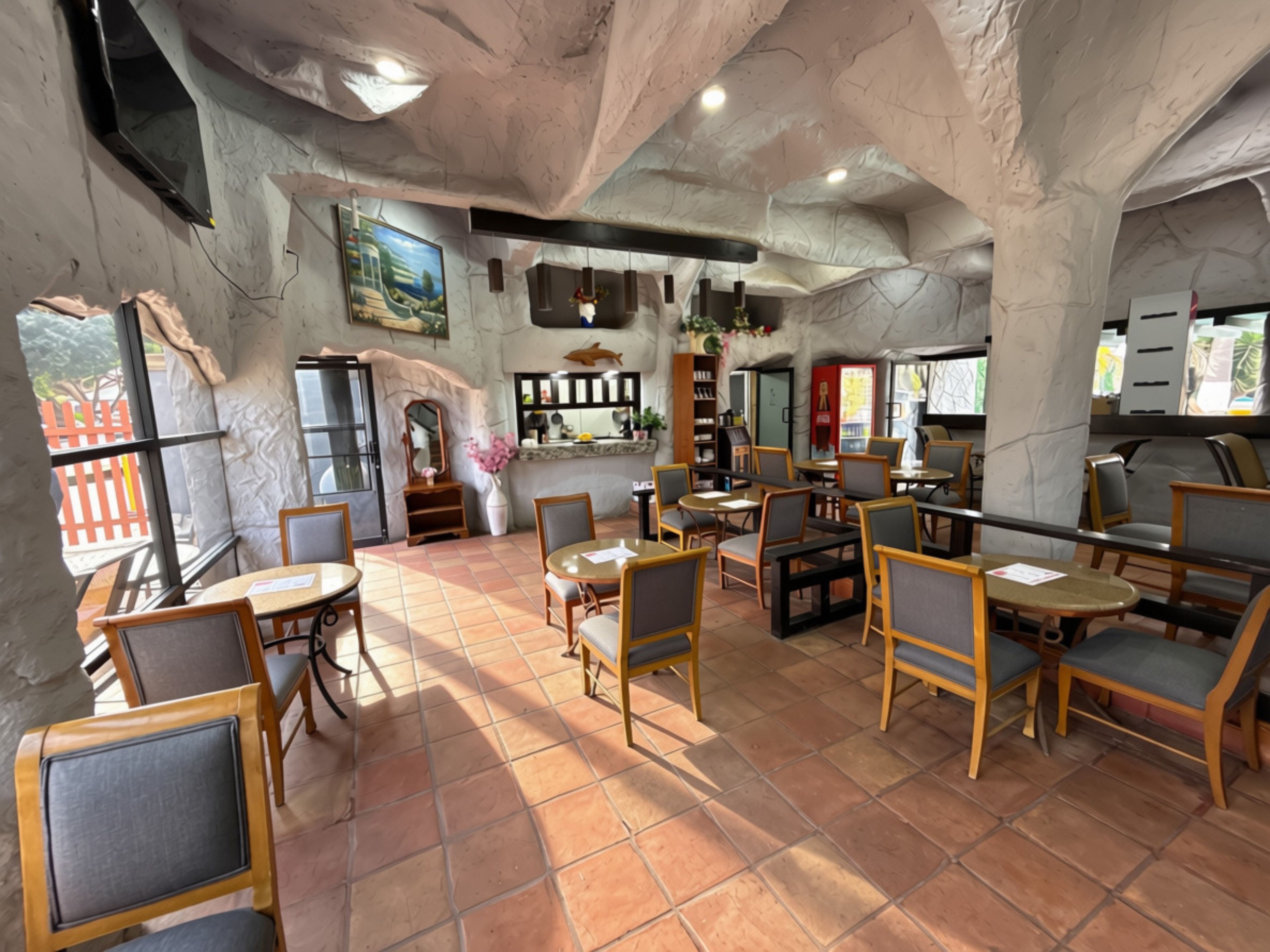 A restaurant interior with tiled floors, wooden tables and chairs, a counter with a display, and a mural on the wall.