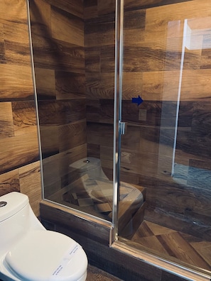 A modern bathroom with a glass-enclosed shower, wooden flooring, and a white toilet. - Hotel Enjoy (Playas de Rosarito)