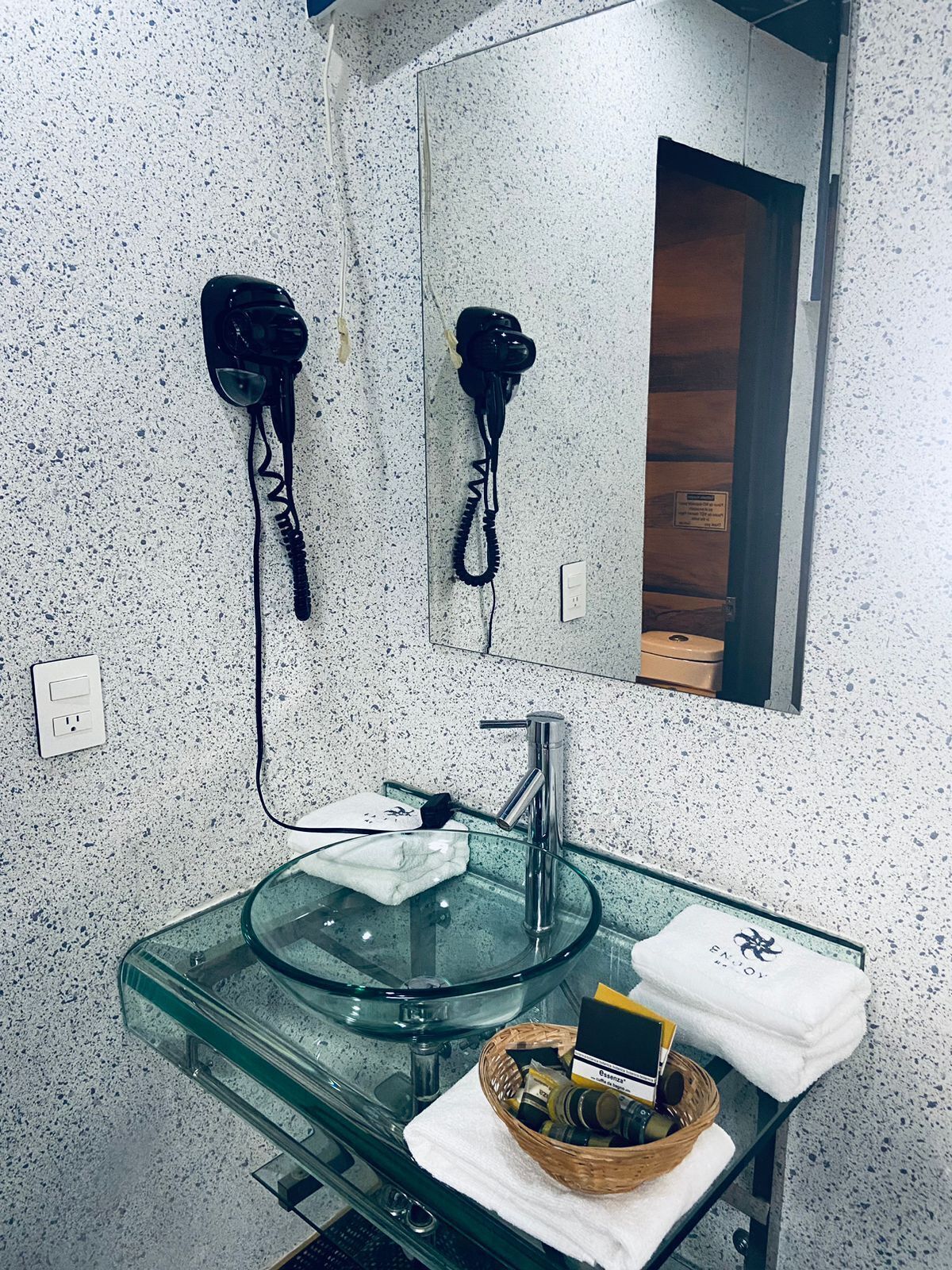 A bathroom with a glass sink, two hair dryers mounted on the wall, a mirror, and a towel rack.