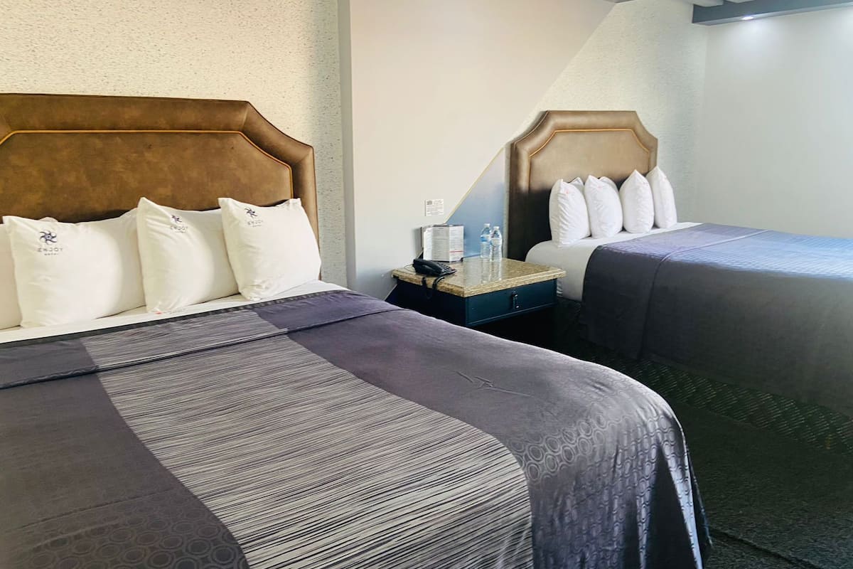 A hotel room with two beds, each with a headboard, a nightstand, and a small table with a lamp and water bottles.