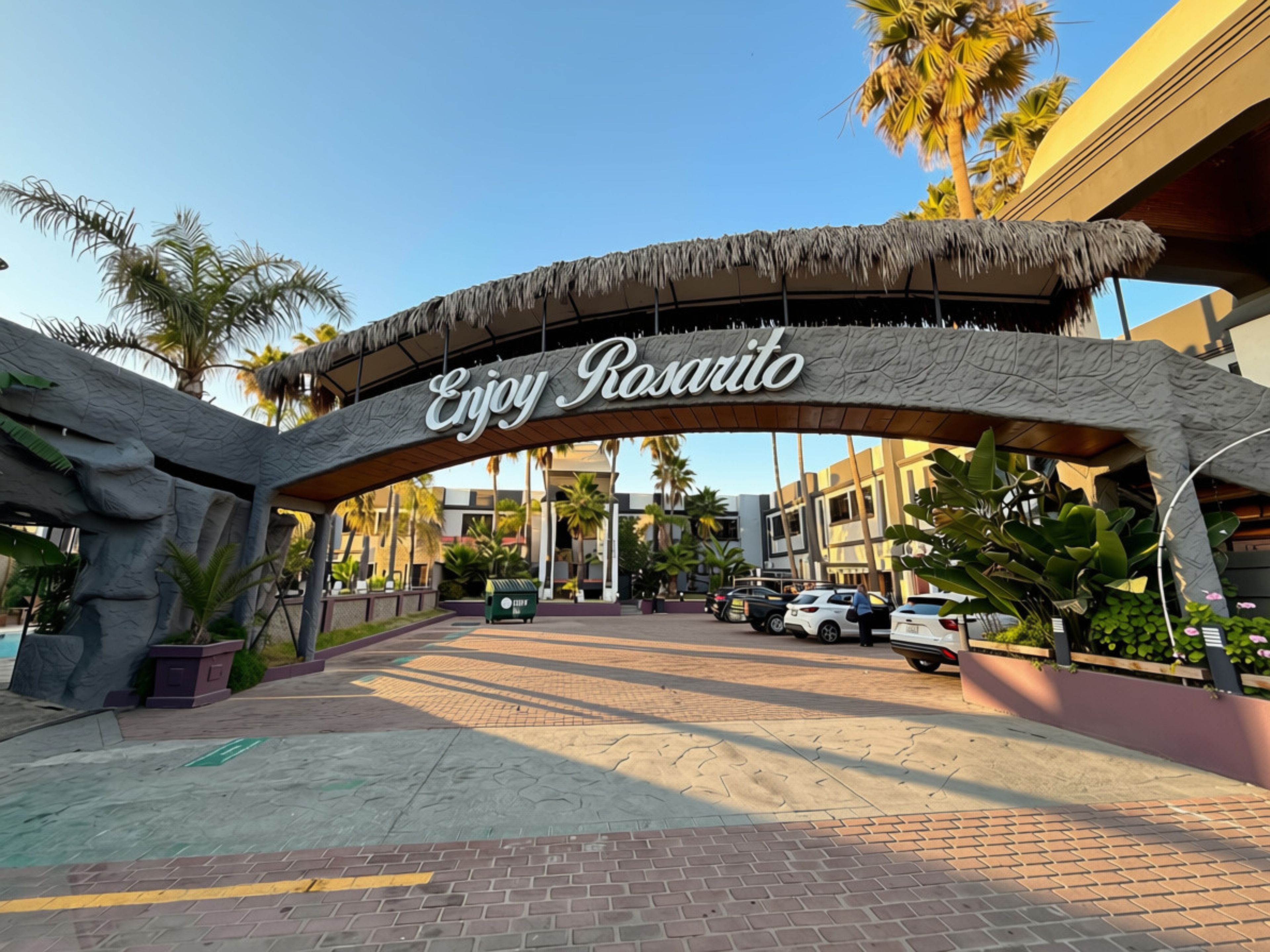 A sign with 'Enjoy Rosarito' written on it, a paved entrance, palm trees, and parked cars.