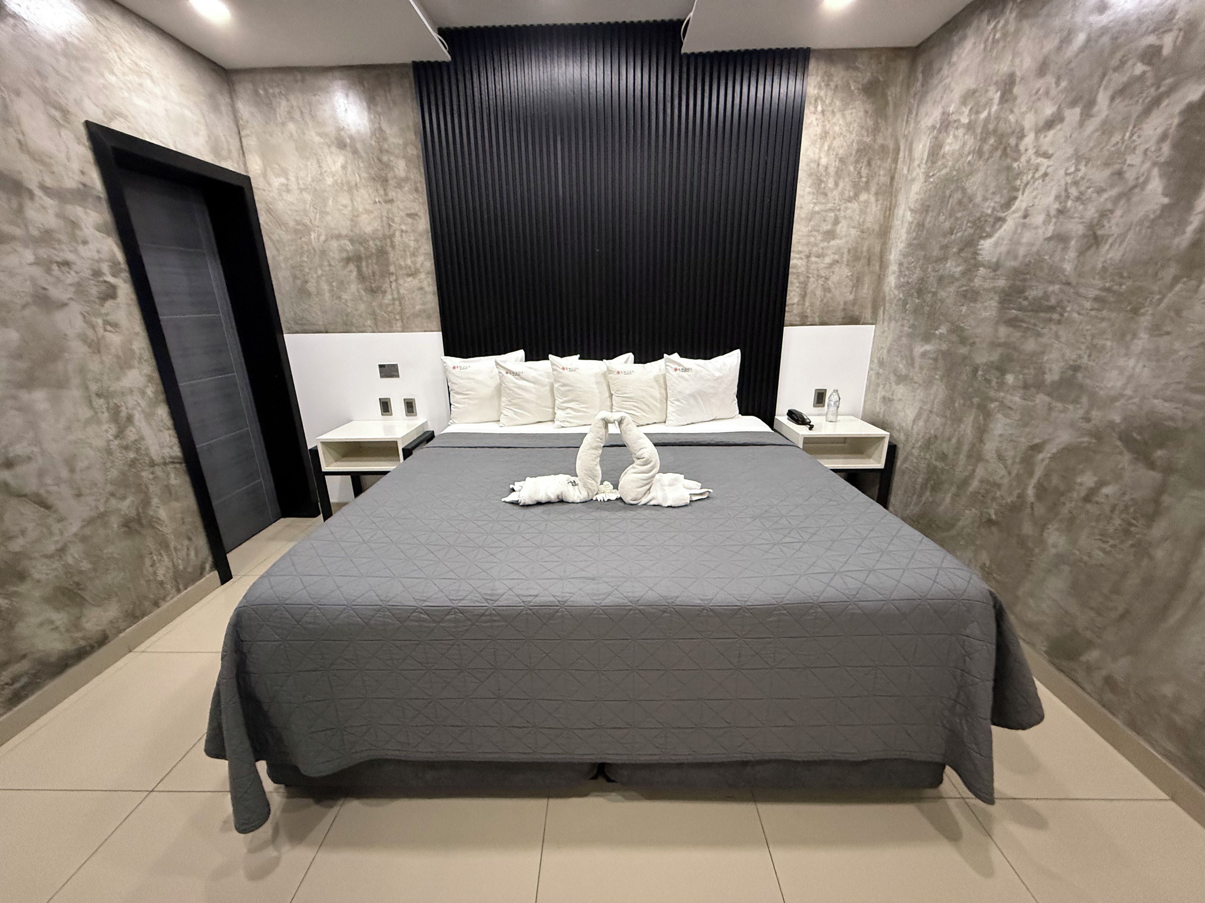 A modern hotel room with a large bed, a headboard, two bedside tables, and a door.
