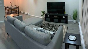 A modern living room with a grey sectional sofa, a black entertainment unit, and a flat-screen TV. - 😍GREAT LOCATION 2BD/1BTH UNIT W/ PVT BACKYARD 😍 (Puyallup)