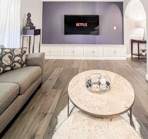 A modern living room with a flat-screen TV mounted on a wall, a grey sofa with patterned cushions, a round coffee table with a glass top, and a wooden floor. - In-Shower Sauna, Fenced Yard W/Deck & BBQ Near ATL (Atlanta)