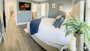 Un dormitorio con una cama grande, un ventilador de techo, un televisor mostrando 'NETFLIX' y un pasillo que da a otra habitación.