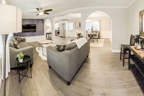 A modern living room with a grey sofa, a coffee table, and a TV. There is a kitchen in the background with a dining area. - In-Shower Sauna, Fenced Yard W/Deck & BBQ Near ATL (Atlanta)
