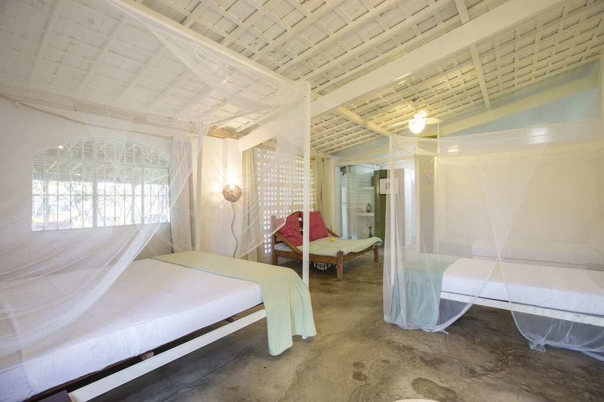 A room with two beds, each covered with mosquito netting, a wooden floor, and a window with a decorative grille.