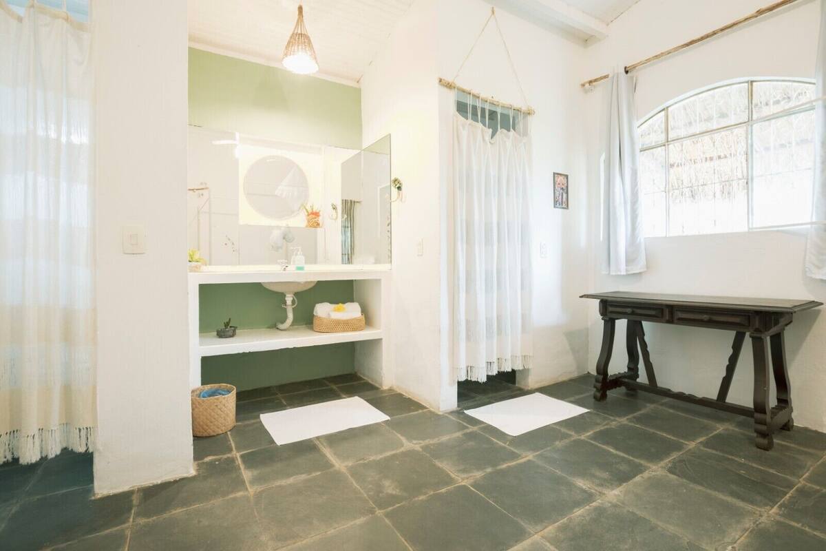 A bathroom with a sink, mirror, and shelves; a window with curtains; and a table with a chair.