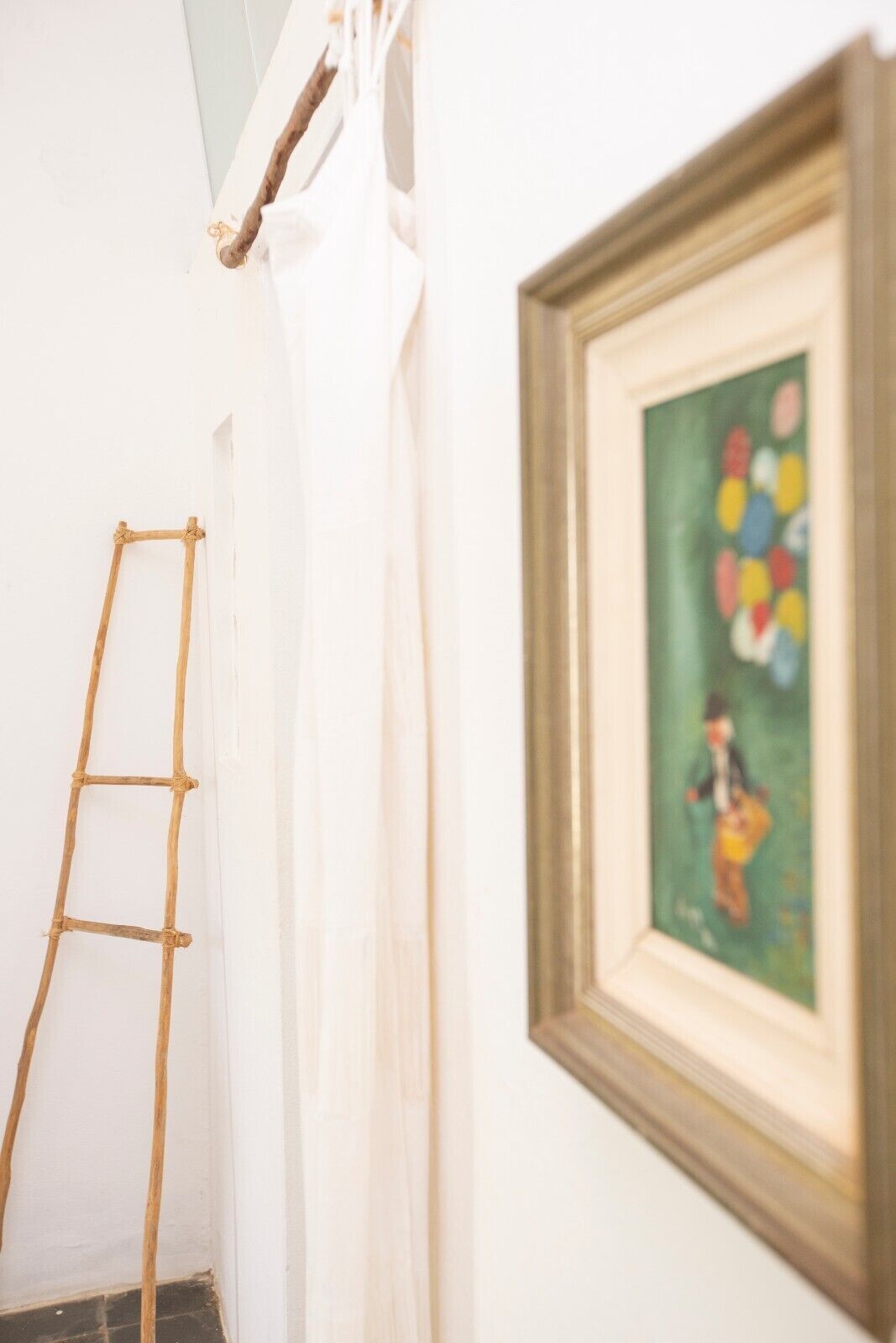 Una escalera apoyada en una pared blanca, un cuadro con fondo verde y una cortina blanca.
