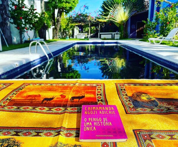 A poolside view with a book titled "CHIMAMANDA NGOZI ADICHIE O PERIGO DE UMA HISTORIA" on a patterned tablecloth. - Villa Ostello Pousada (Paripueira)