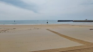 Uma extensa praia de areia com um farol ao longe e uma estrutura de quebra-mar.