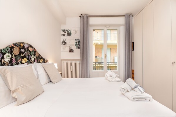 Habitación de hotel con cama, radiador, ventana con cortinas y vista al balcón exterior.