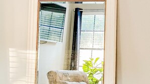 Un grand miroir à cadre blanc reflète une pièce où l’on aperçoit un lit, une fenêtre avec des stores et une plante.