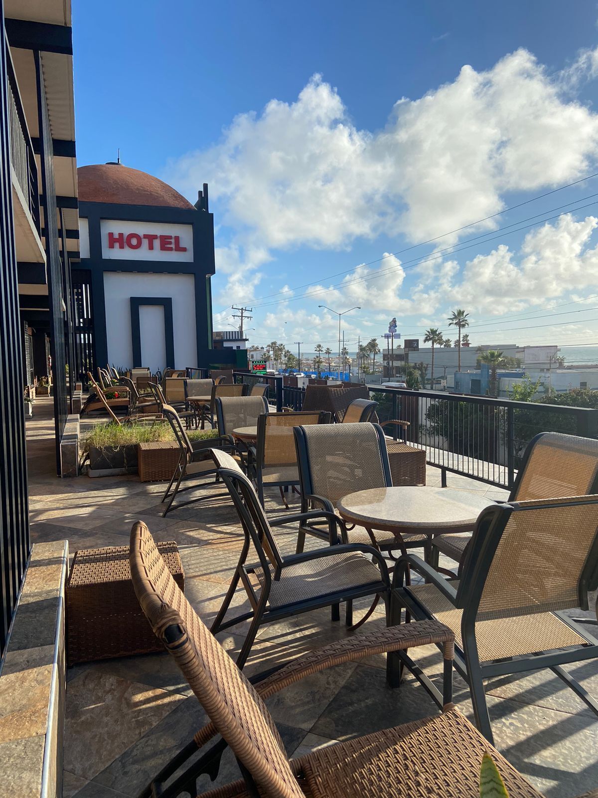 Área de estar externa com cadeiras e mesas de vime, com vista para um horizonte urbano que inclui a placa de um hotel.