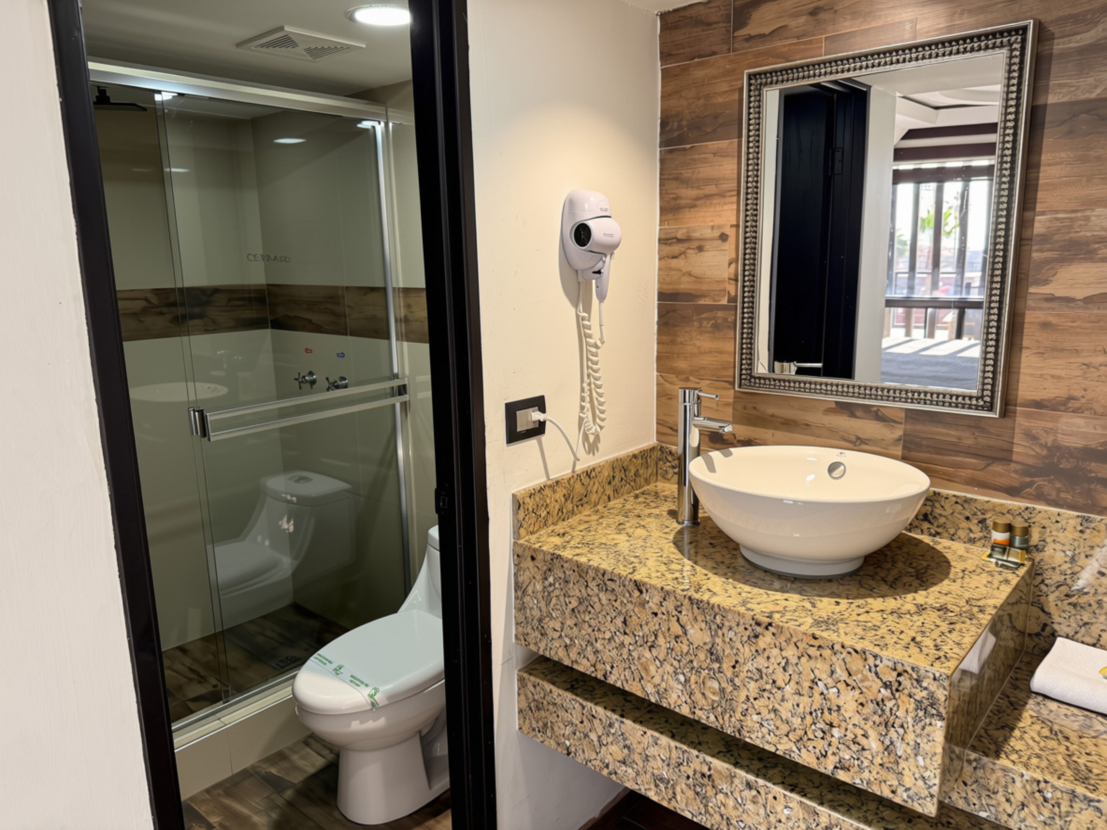 Un baño con ducha acristalada, inodoro blanco, lavabo con encimera de mármol y espejo con marco decorativo.