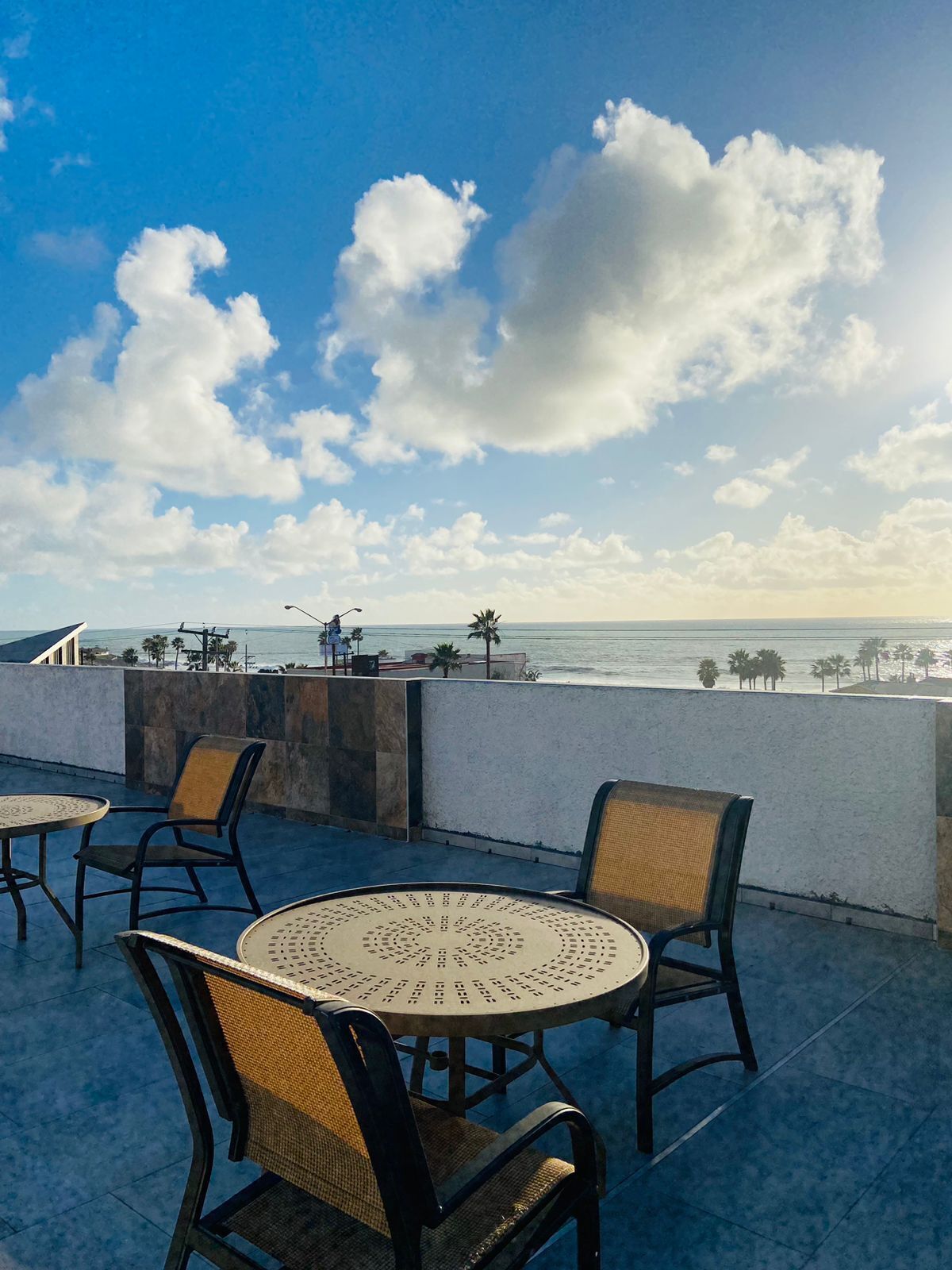 Terraço com mesas e cadeiras redondas, com vista para a costa, palmeiras e céu claro.
