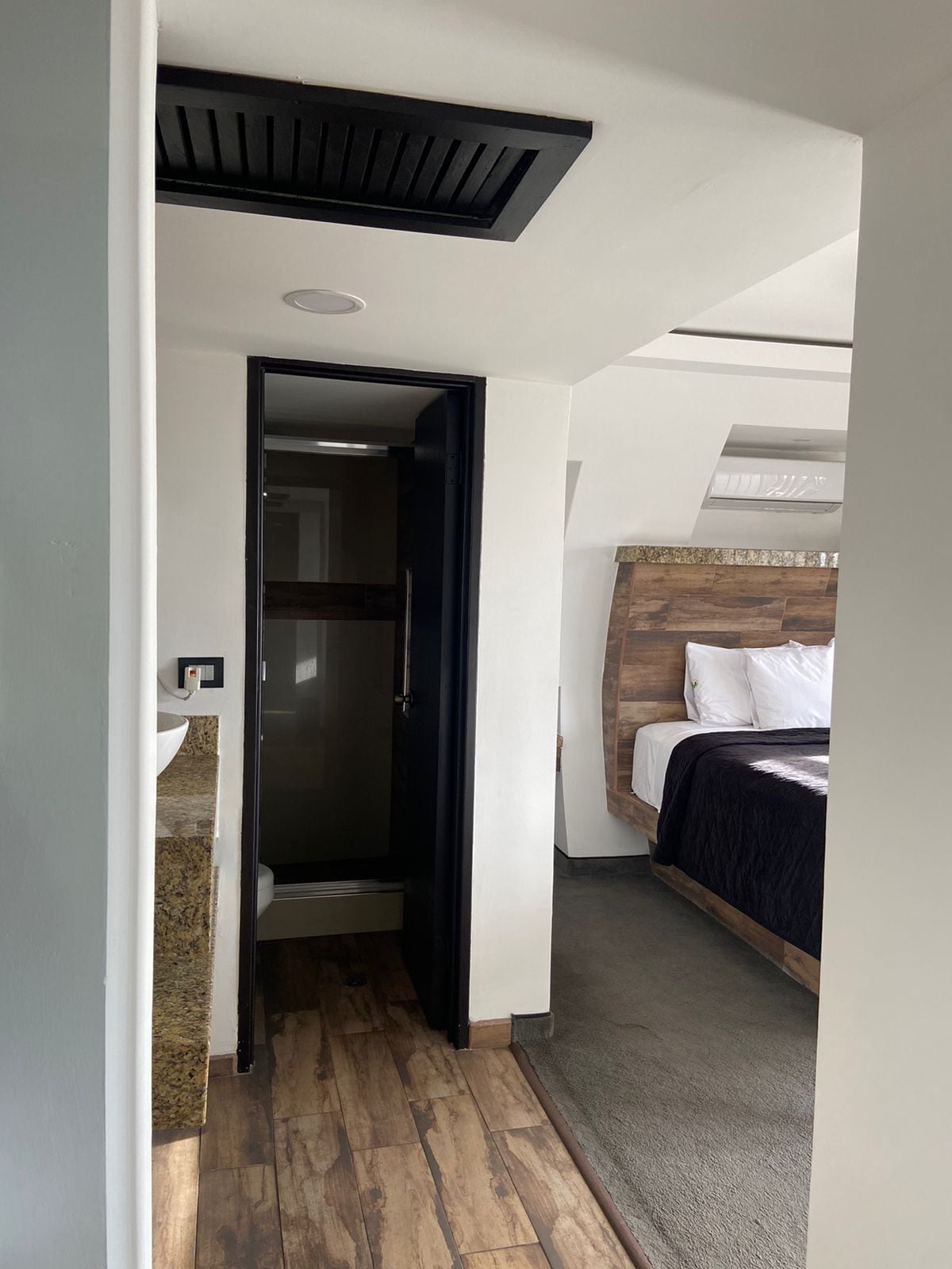 A hotel room with a wooden headboard, a bed with a dark cover, a built-in heater, and a small bathroom with a sink and mirror.