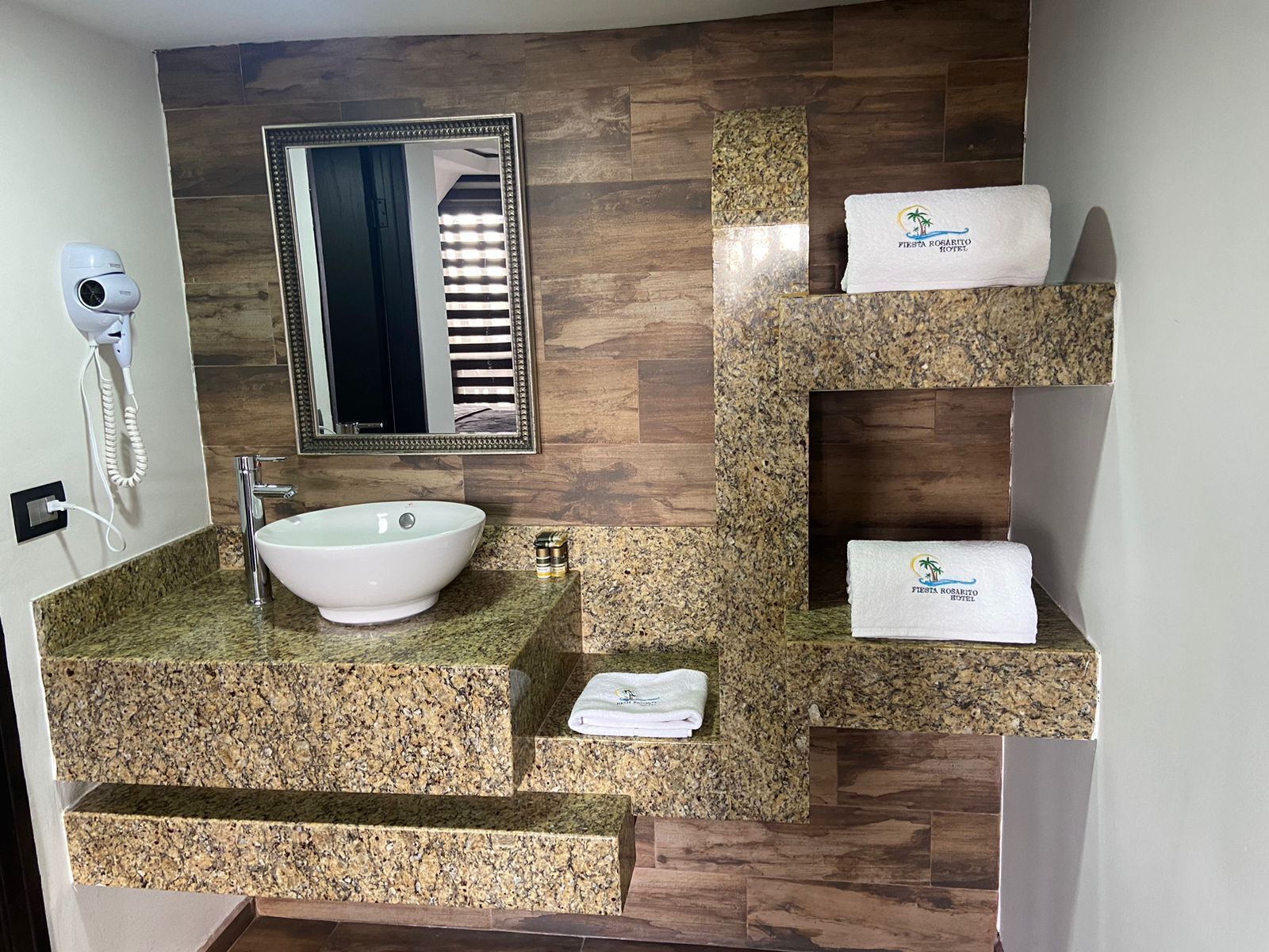 A bathroom with a granite countertop, a white sink, a mirror, a hairdryer, and two towels with the hotel logo.