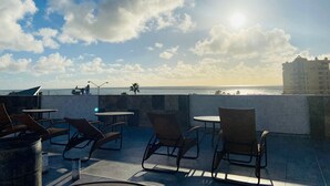 Una terraza en la azotea con mobiliario exterior, vista al océano y un edificio lejano.