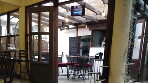 Un interior con zona de comedor con mesas y sillas, un televisor montado en la pared y piso de baldosas.