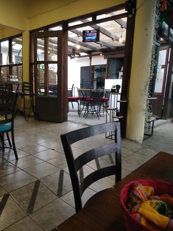 Un interior con zona de comedor con mesas y sillas, un televisor montado en la pared y piso de baldosas.