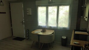 A small, compact room with a round table, a window with curtains, a door, and a kitchenette area.