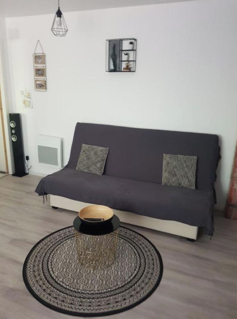 Un salon moderne avec un canapé gris foncé, deux coussins à motifs, une table basse ronde dont le plateau est en matière tressée, et une étagère murale agrémentée d’objets décoratifs.
