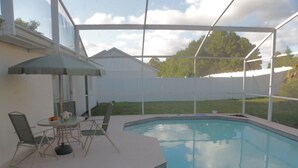 Piscina exterior techada con mesa y sillas, rodeada por una valla blanca y un césped.