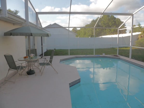 Et overdækket udendørs swimmingpool med borde og stole, omgivet af et hvidt hegn og en græsplæne.