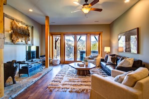 A spacious living room with a large window, a ceiling fan, a fireplace, and a stone fireplace surround.