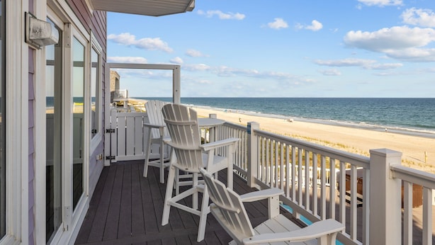 A wooden deck with white Adirondack chairs and a table, overlooking a beach with a clear sky. - The Pinnacle - OCEANFRONT Home.  Amazing Views, Pool, Hot Tub, 2 Kitchens,
Deck (Emerald Isle)