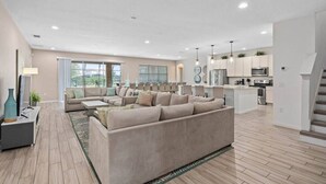 A modern living room with a large beige sectional sofa, a coffee table, and a TV. The room has a large window with a view of greenery. There is a kitchen area with white cabinets and stainless steel appliances. - Huge, Popular Resort Villa 30+ Guests - Resort/Pool/Spa/Game Room/ Near Disney! (Davenport)