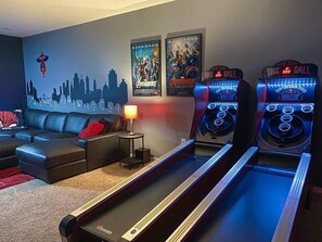 A living room with a sofa, a lamp, two arcade machines, and posters on the wall.