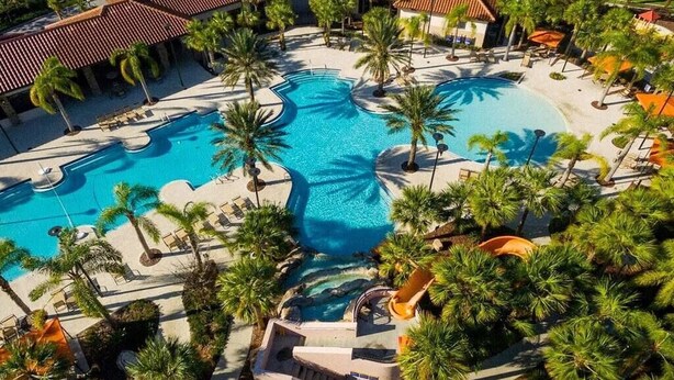 A large outdoor swimming pool surrounded by palm trees and lounge chairs, with a building in the background. - Huge, Popular Resort Villa 30+ Guests - Resort/Pool/Spa/Game Room/ Near Disney! (Davenport)