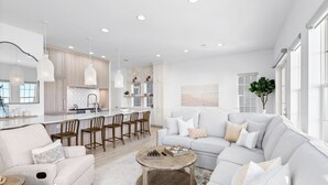 A modern living room with a sofa, coffee table, and chairs, a kitchen with wooden cabinets and pendant lights, and a large window providing natural light. - 🌅 16 Sunsets | Gulf Front | Private Pool | Free Sunset Dolphin Cruise | Beach Chair Service 🐬 (Miramar Beach)