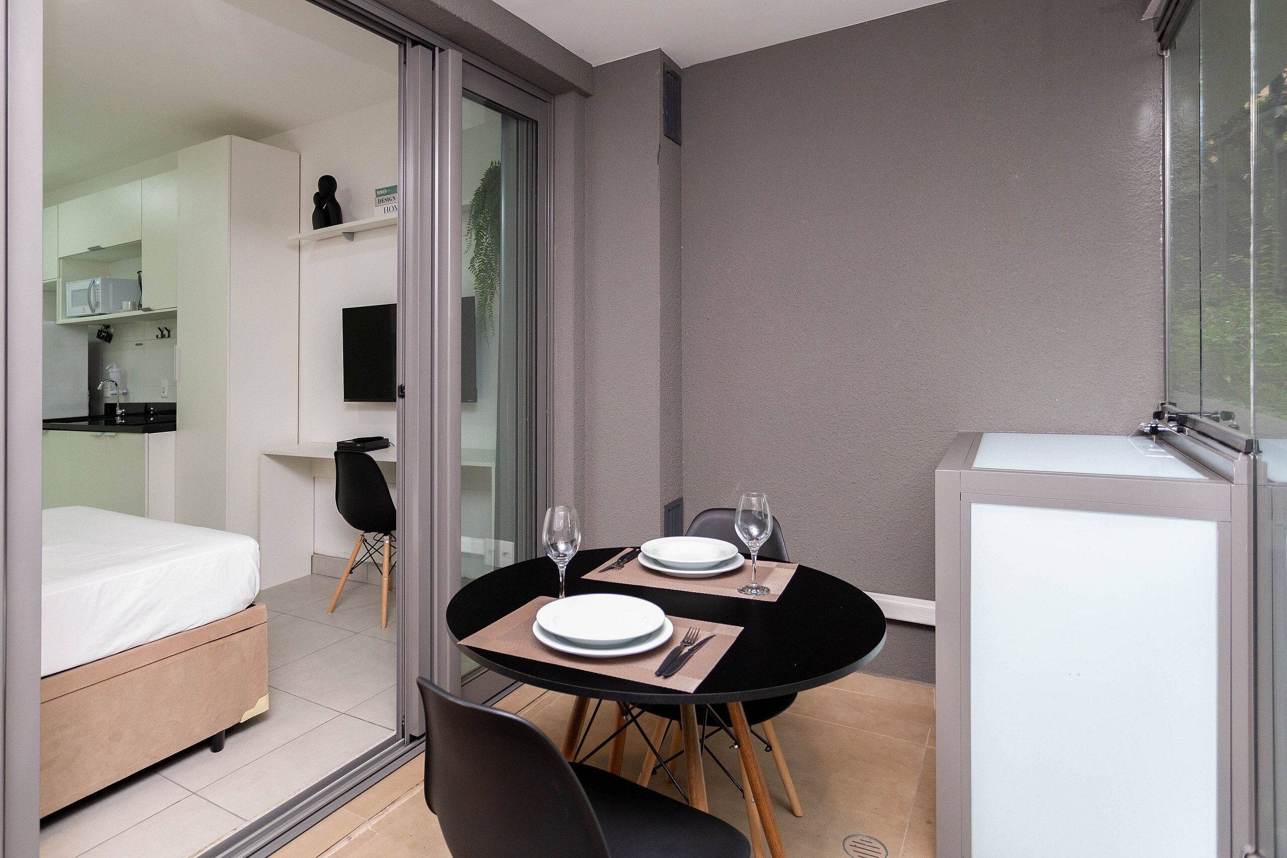 Una habitación compacta con una cama, una pequeña mesa de comedor con sillas, un refrigerador y una puerta corrediza de vidrio que da a un balcón.