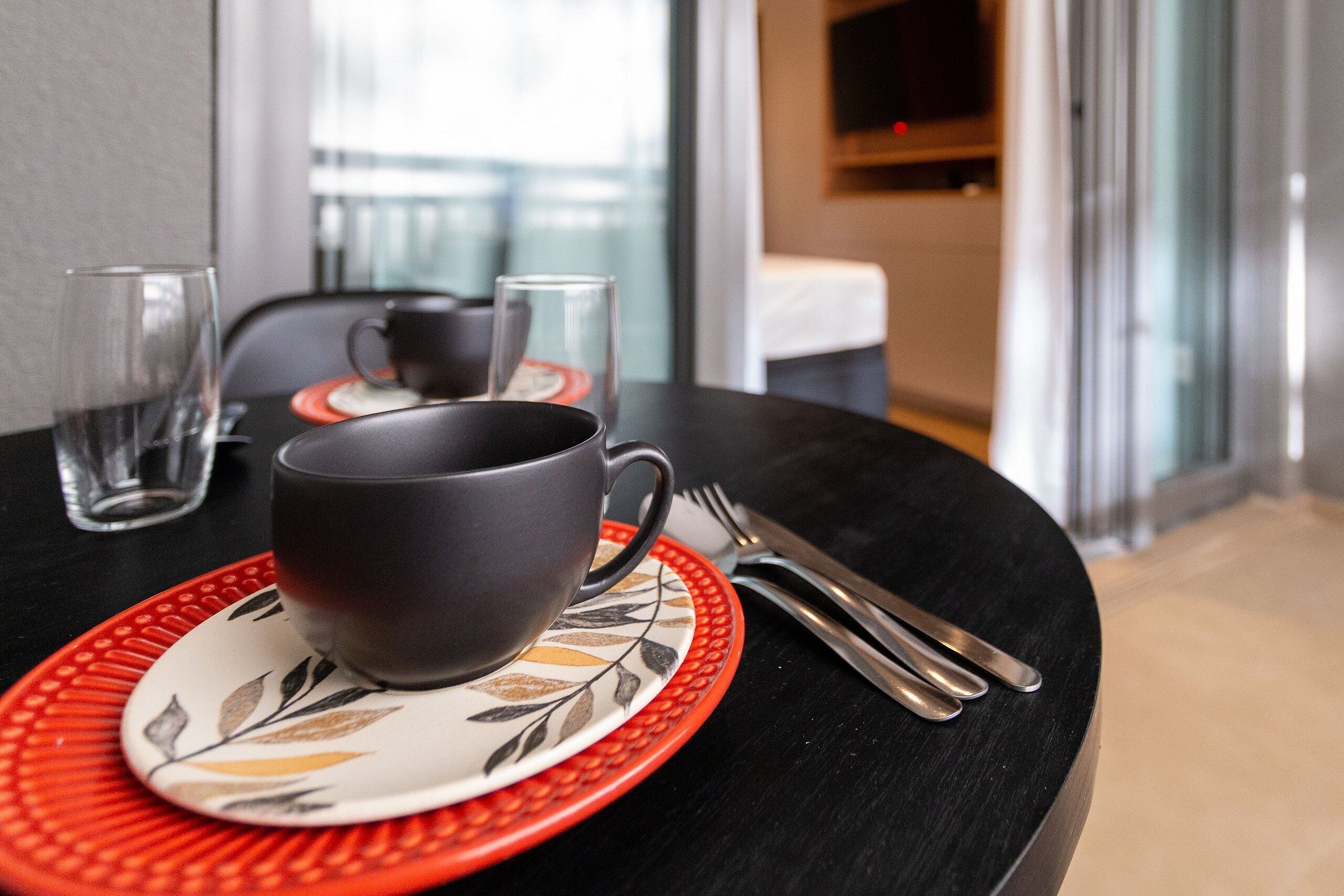 Una mesa con una taza negra, un vaso y un tenedor sobre un mantel individual rojo.