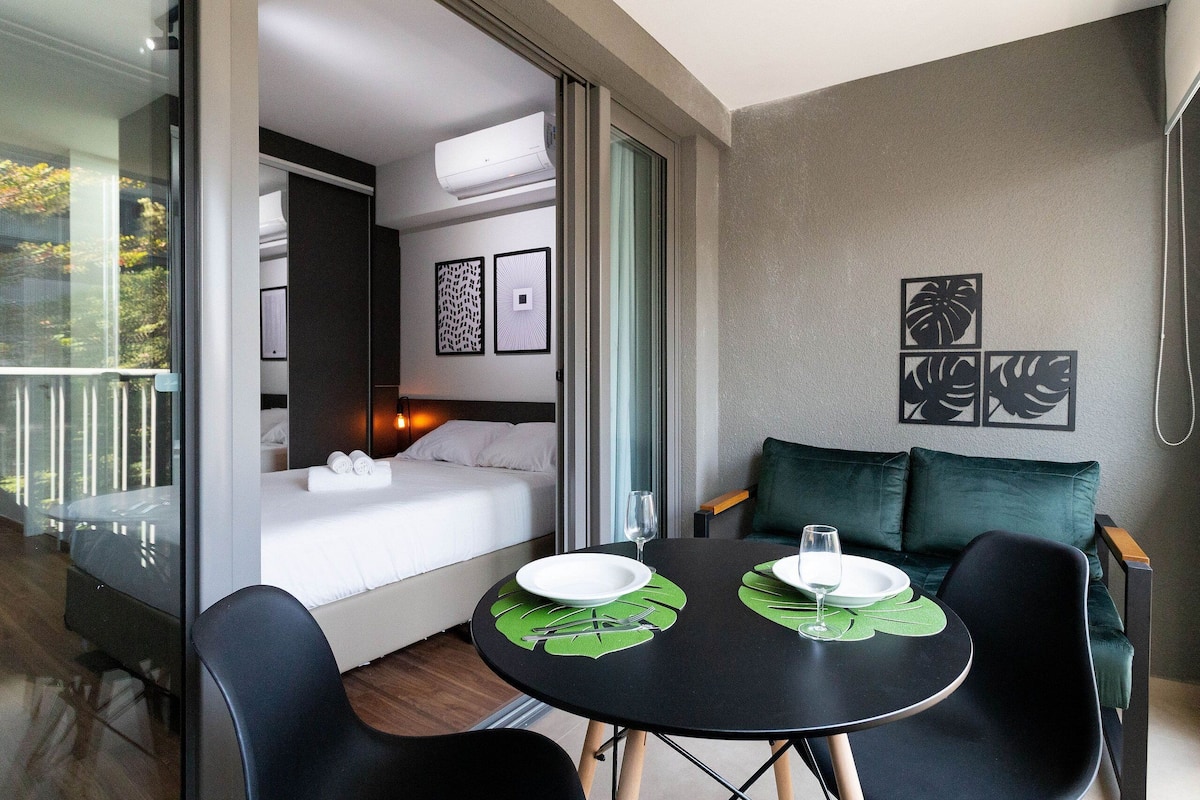 Una habitación de hotel moderna con balcón, cama con ropa blanca, una mesa de comedor con dos sillas y un sofá verde.