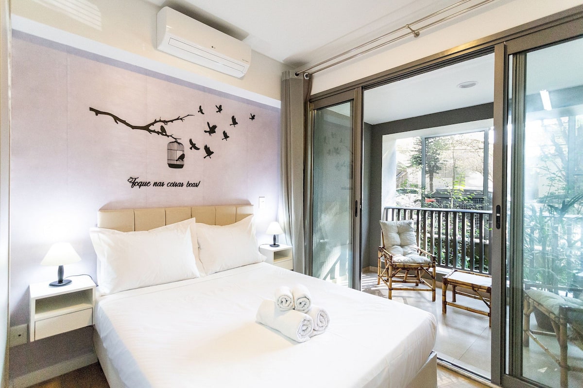 Habitación de hotel con cama, mesita de noche, decoración en la pared y balcón con silla y mesa.