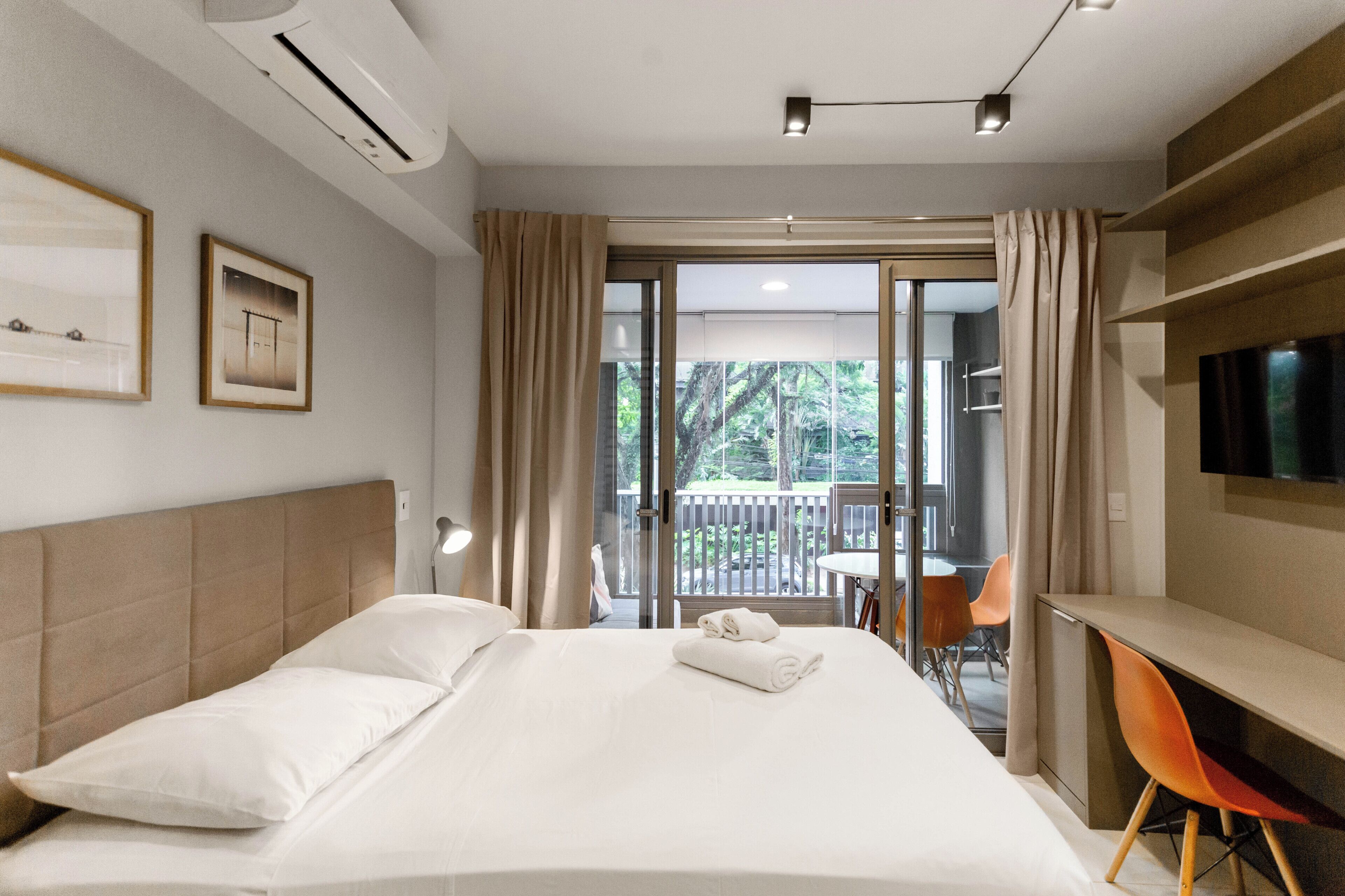 Habitación de hotel con cama, escritorio, silla, televisor y balcón con vista a árboles.