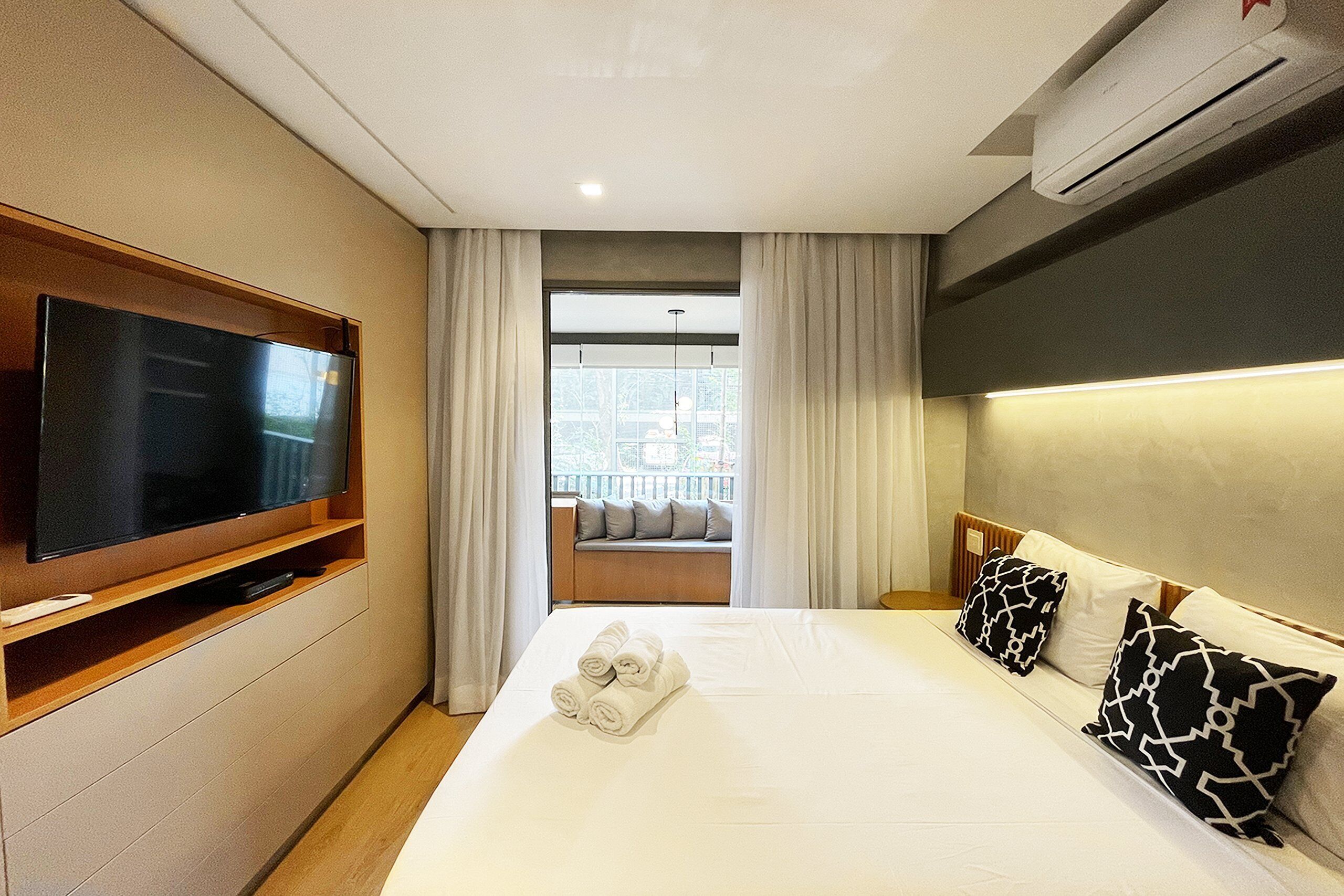 A hotel room with a large bed, a flat-screen TV mounted on the wall, a window with curtains, and a view of the city.