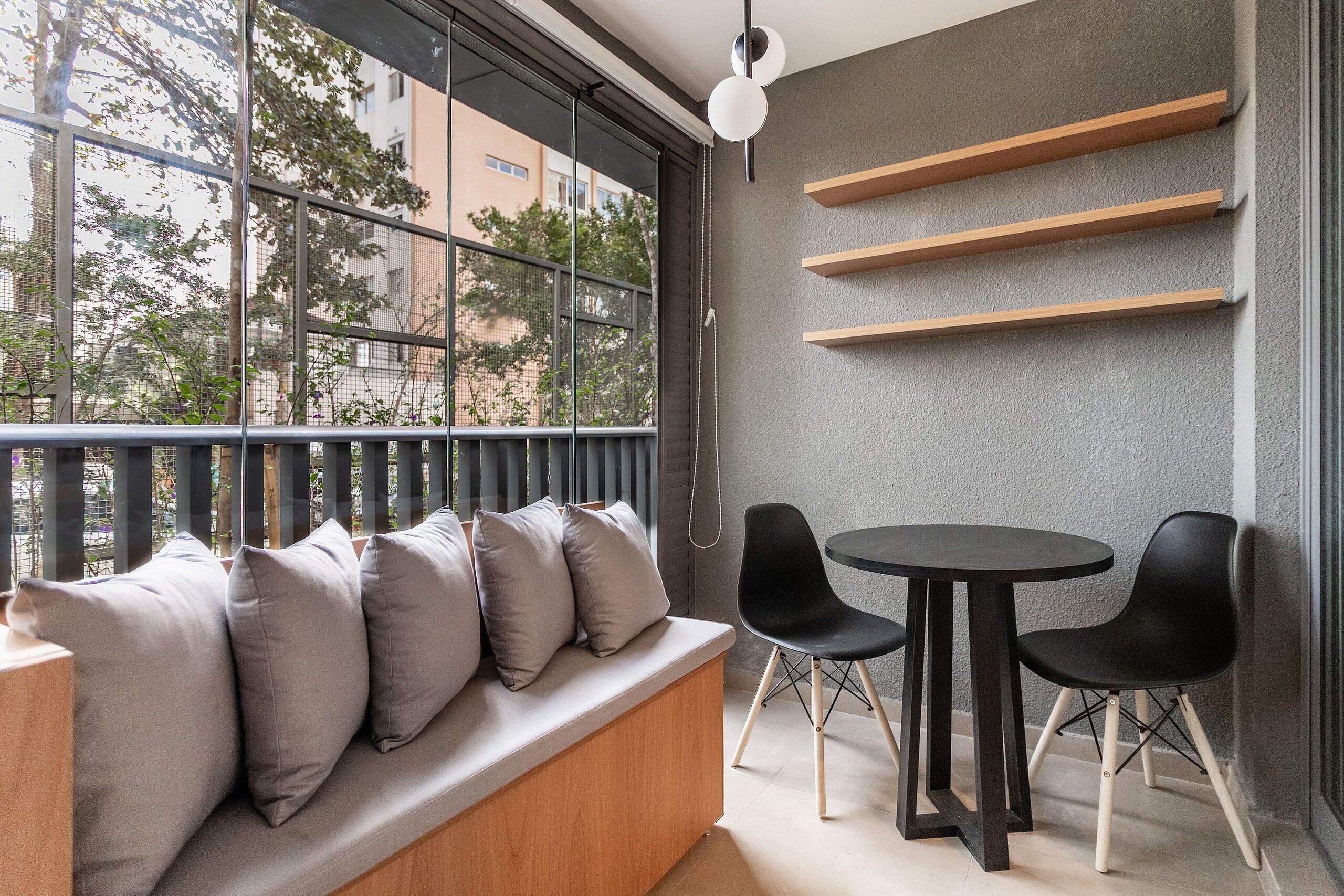 A modern balcony with a wooden bench and black chairs, a grey textured wall, and a view of buildings and trees.