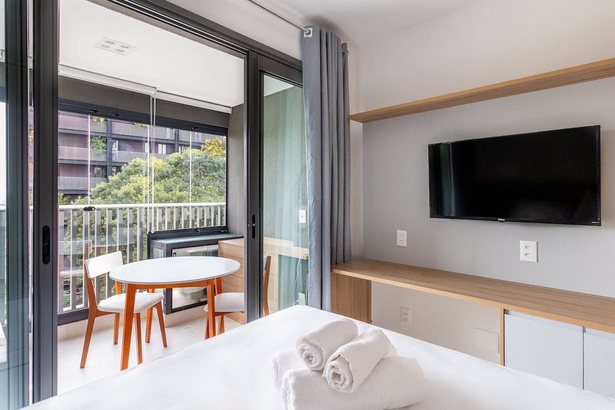 Habitación de hotel con cama, televisión, balcón con mesa y sillas, y vistas a edificios y árboles.