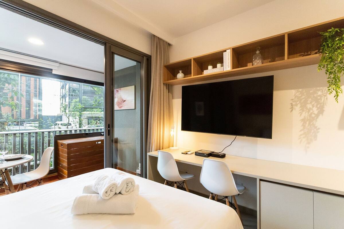 Habitación de hotel con cama, dos sillas blancas, un escritorio, televisor y balcón con vista a edificios y árboles.
