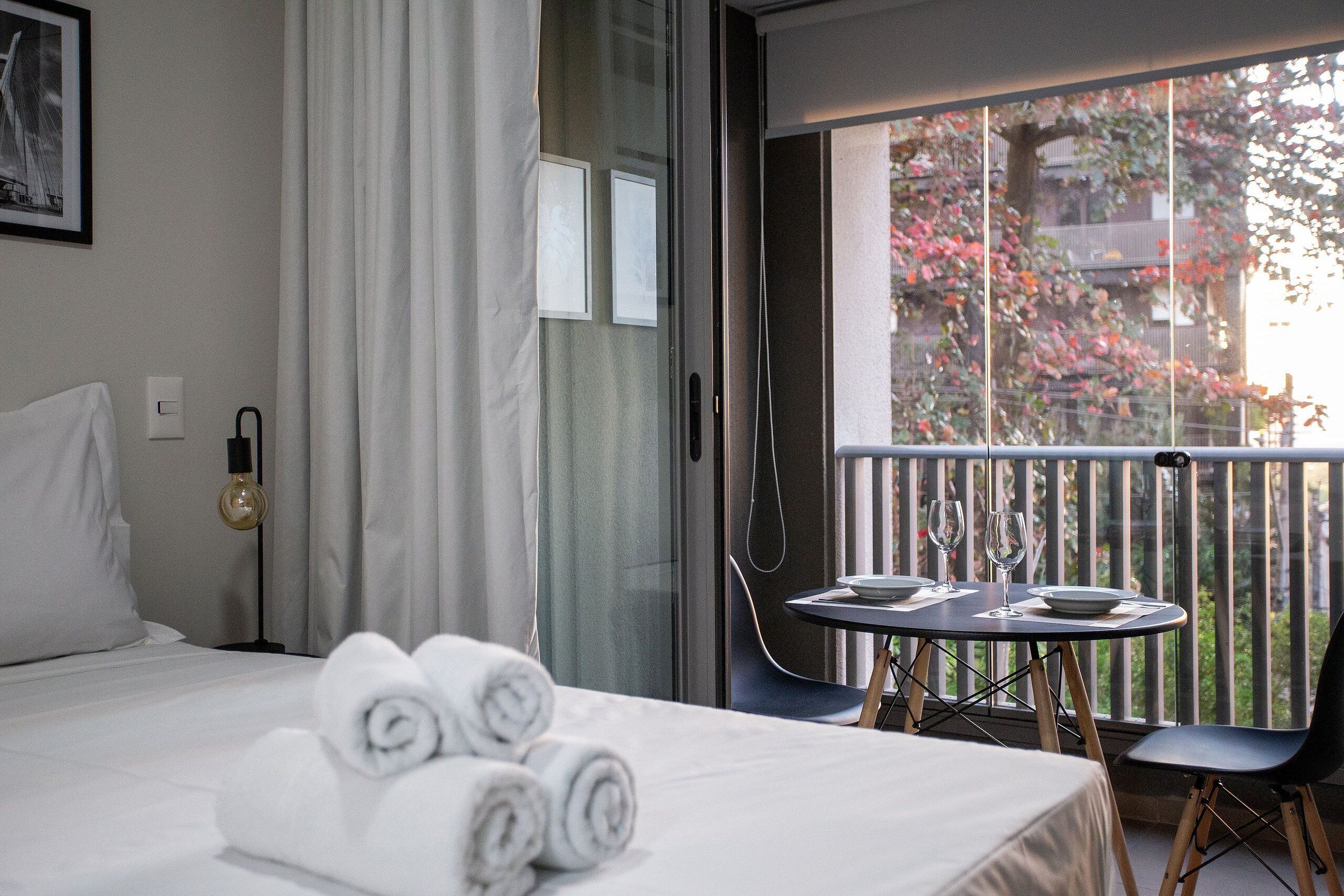 Une chambre d’hôtel comprenant un lit, une table avec une chaise et un balcon donnant sur des arbres et des immeubles.