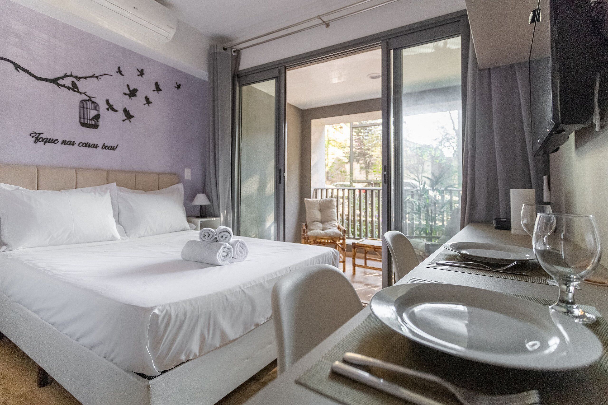 A hotel room with a bed, a dining area with a table set for two, and a balcony with a chair.